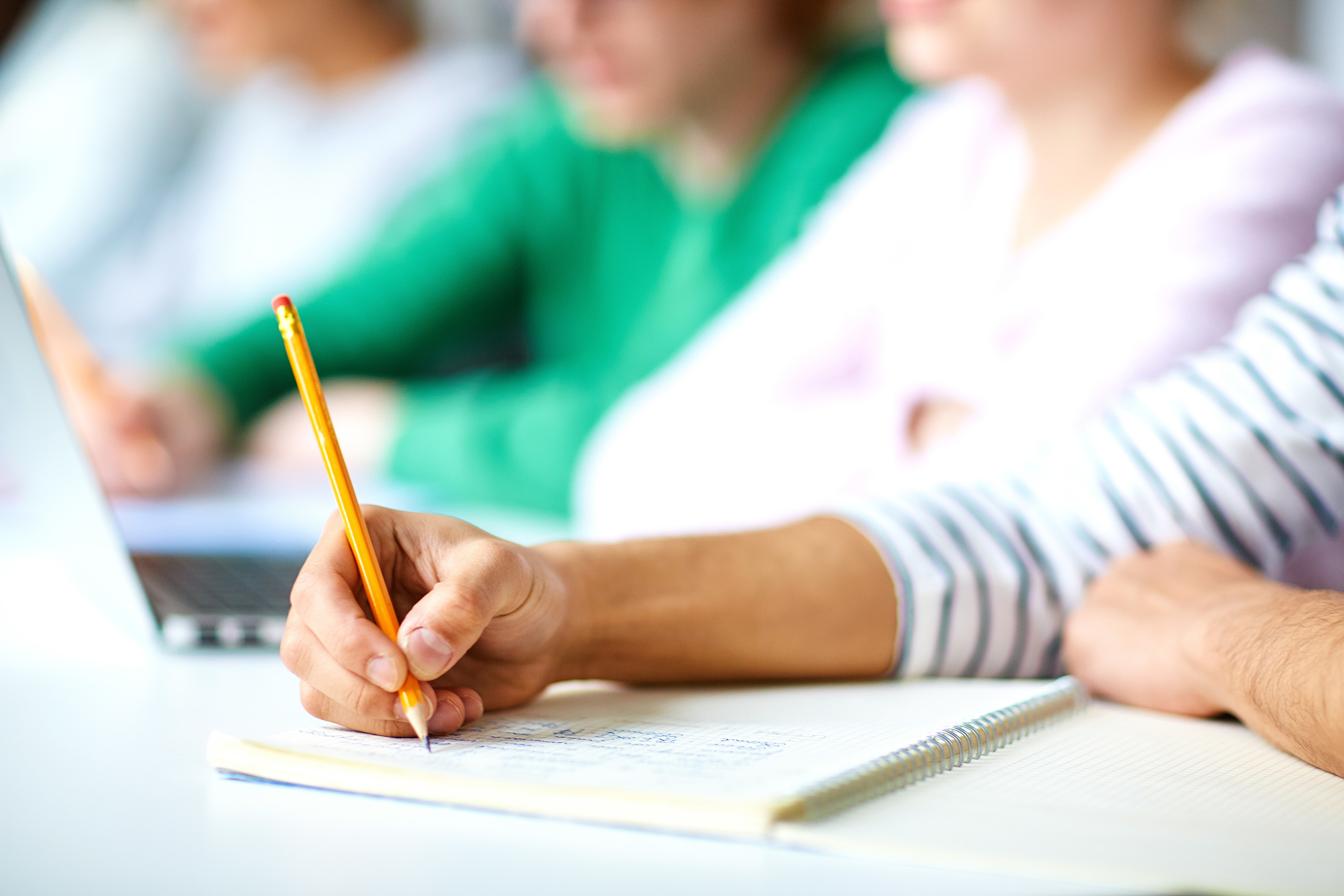 Eine Hand hält einen Bleistift und schreibt in ein Notizbuch, im Hintergrund eine Gruppe, die an einem Lern- oder Coaching-Setting teilnimmt – passend zu Themen wie mentale Stärke und persönliche Leistungsfähigkeit.